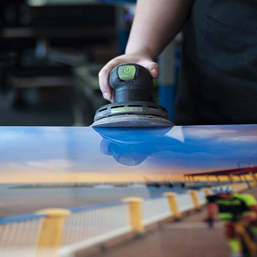 Person using a sander on a reflective metal surface with a blurred outdoor scene in the background
