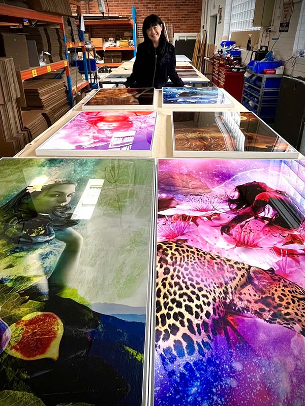 Linda standing behind a table displaying various colorful metal artworks in the Print 2 Metal workshop