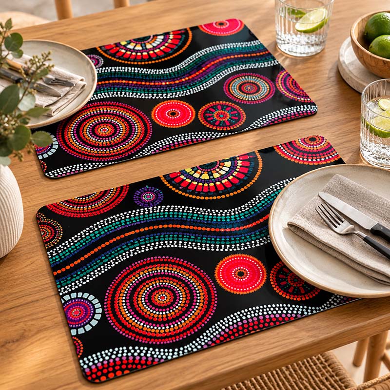 Two rectangular placemats with a vibrant Aboriginal dot art pattern on a black background, styled on a wooden dining table with plates, cutlery, and glasses.